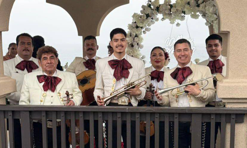 Adult mariachi group smiling and holding instruments, dressed in traditional white attire with burgundy bow ties.
