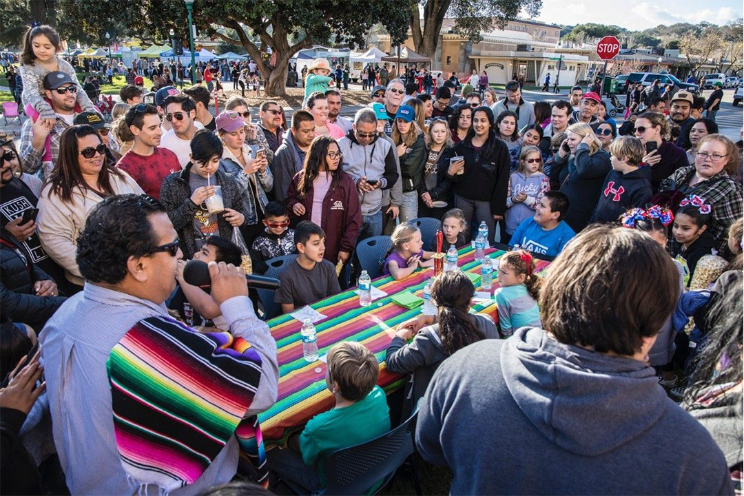Image of kids at a long table preparing for a Tamale Eating Contest, with the event Mc and crowd standing by. - Photo by Rick Evans